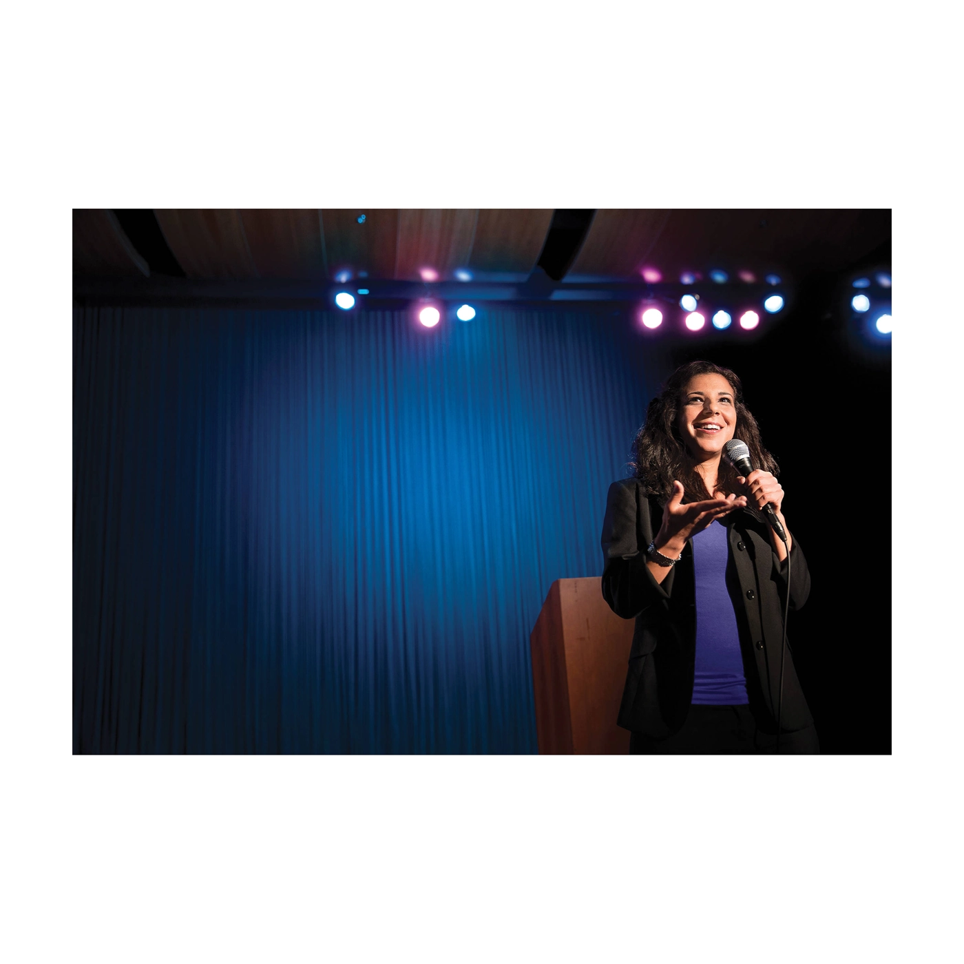 Woman on stage holding a microphone with blue curtains and lights in the background